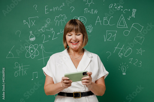 Fun teacher mature elderly woman 55 wear white shirt using play racing app on mobile cell phone hold gadget smartphone for pc video games isolated on green wall chalk blackboard background studio. © ViDi Studio