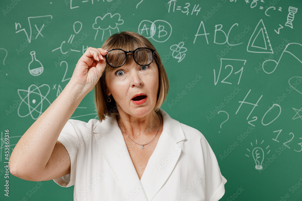 Shocked teacher mature elderly lady woman 55 wear shirt raised glasses keeping mouth wide open isolated on green wall chalk blackboard background studio. Education in high school September 1 concept.