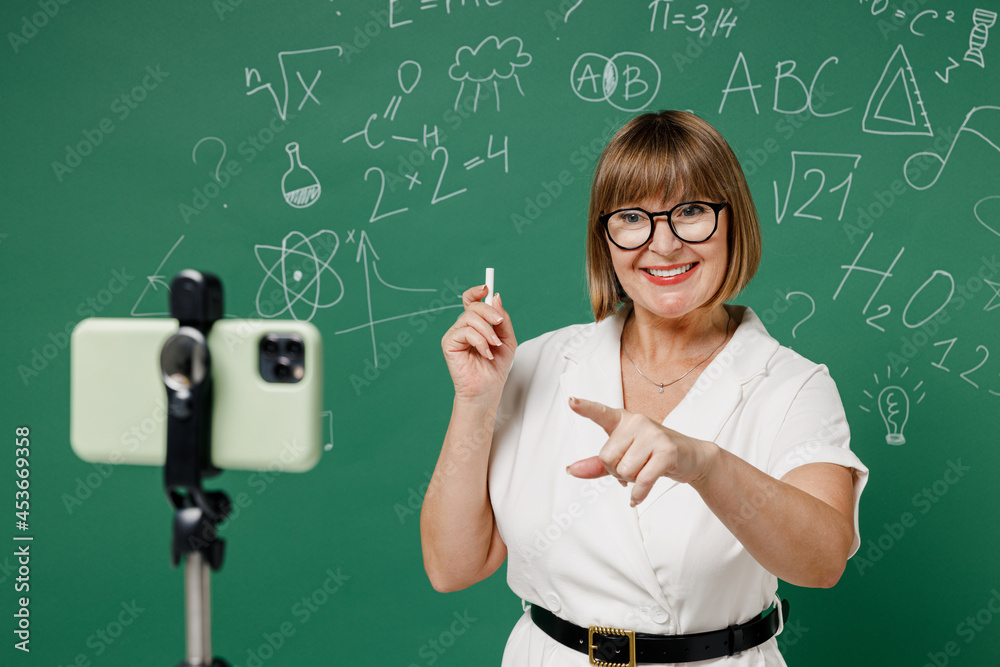 Teacher mature elderly lady woman 55 wear shirt glasses work with mobile cell phone on tripod on lockdown quarantine point index fingers camera isolated green wall chalk blackboard background studio.