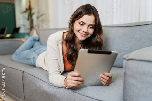 Happy Latin lady relax at home alone on room pose share good news at social media via digital tablet. Smiling woman enjoy weekend order goods food online in tablet app.