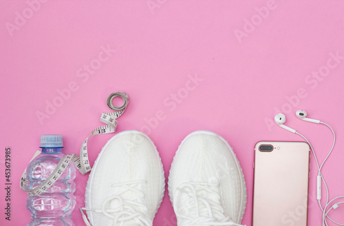 A fitness concept with a water bottle, sneakers, a phone with headphones, a pink roller on a pink background. A place to copy.