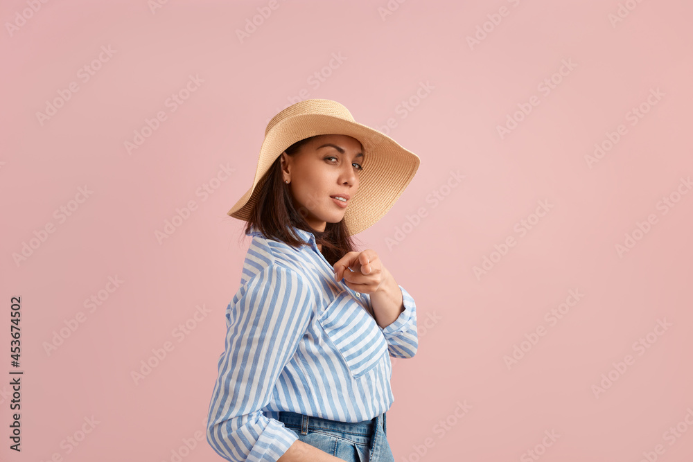 Sexy woman with dark hair makes shooting gesture at camera, wants you, poses in straw hat, flirts, feels sexy, wears striped shirt, jeans with belt, on pink background. Summer emotions concept. Sexy woman with dark hair makes shooting gesture at camera, wants you, poses in straw hat, flirts, feels sexy, wears striped shirt, jeans with belt, on pink background. Summer emotions concept.
