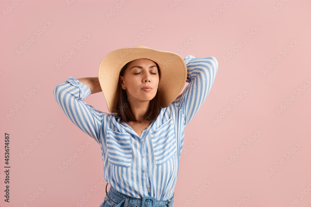 Relaxed brunette enjoys free time on the beach, closes eyes, dreams of vacation, holds hands behind head, wears straw hat, striped shirt, jeans with belt, on pink background. Summer emotions concept. Relaxed brunette enjoys free time on the beach, closes eyes, dreams of vacation, holds hands behind head, wears straw hat, striped shirt, jeans with belt, on pink background. Summer emotions concept.