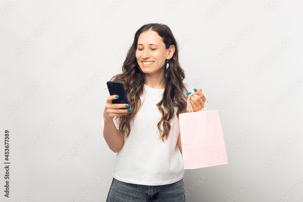 Beautiful young lady is holding a shopping bag, while ordering something or paying by phone. Photo made in a white studio. Beautiful young lady is holding a shopping bag, while ordering something or paying by phone. Photo made in a white studio.