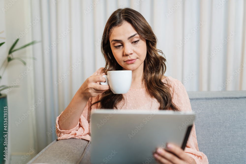 Happy Latin lady relax drinking coffee at home alone sit on room using digital tablet.