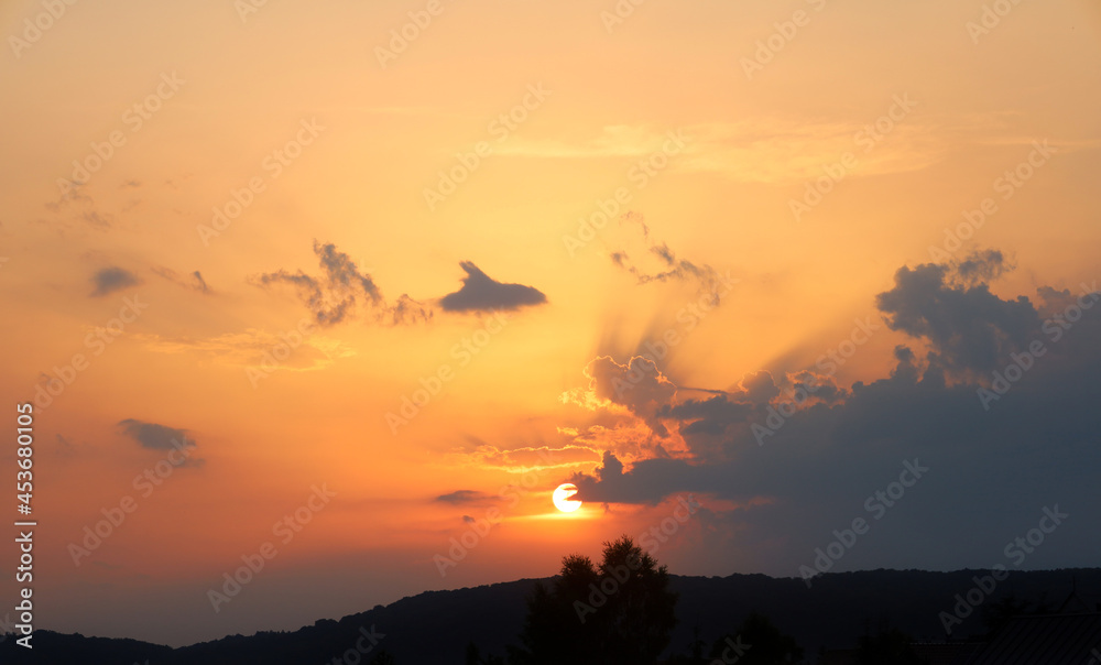 Sunset time over silhouettes of hills covered with forest and trees. Orange sky and big dark purple cloud that partially obscured the sun's bright disk. Diverging rays from the sun.