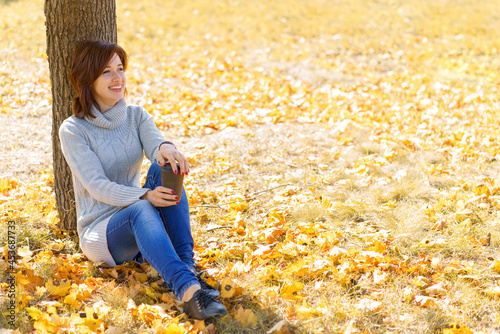 Beautiful young brunette sits on fallen autumn leaves in park  leaning her back against a tree trunk. Girl in a knitted gray sweater. Autumn sunny day in the park.