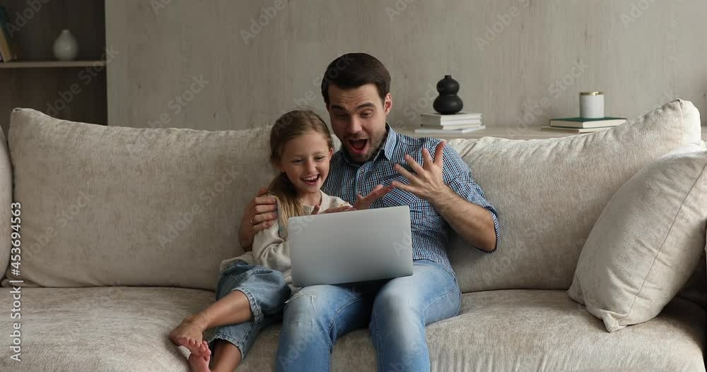 Young father and little adorable 8s daughter sit on comfy couch at living room using laptop enjoy weekend free time on internet, watching together cartoons on-line, enjoy video-sharing website content