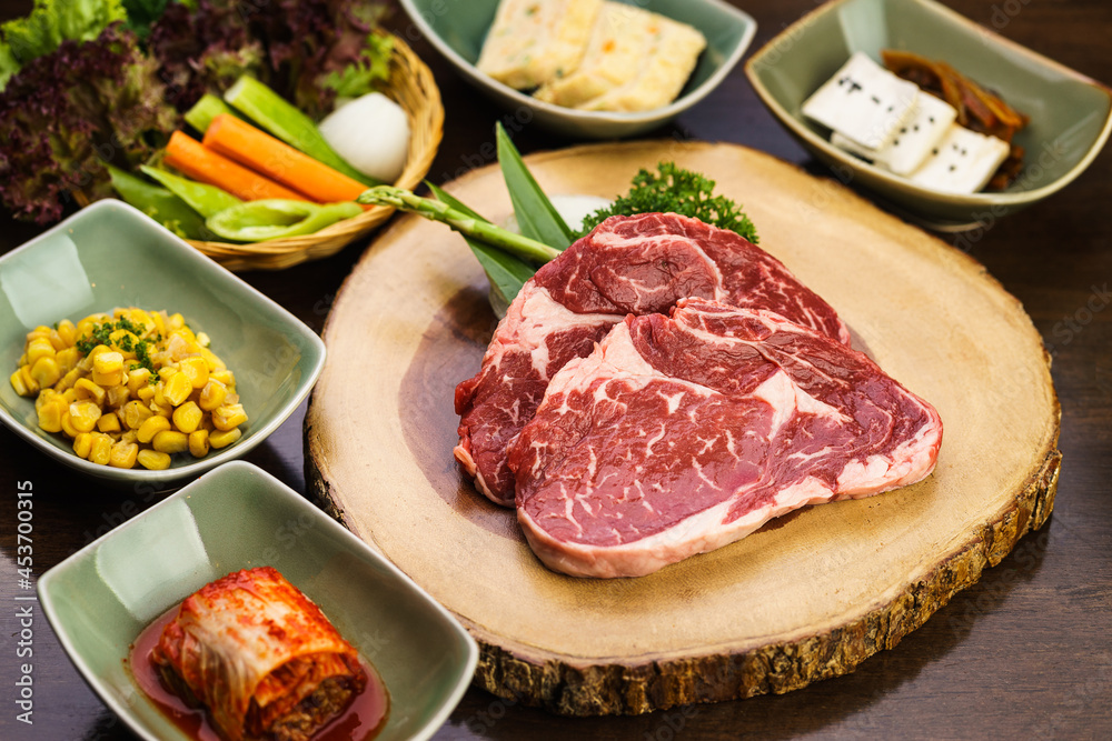 Fresh and raw expensive A5 Wagyu marble beef meat ready to grill on a wooden tray with kimchi side dish and vegetable. Traditional Korean BBQ or Japanese Yakiniku Barbecue concept. Selective focus.