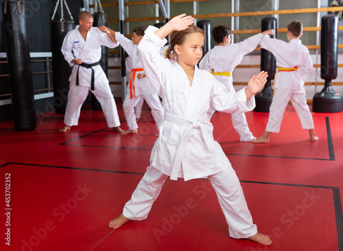 Adolescent girl in kimono posing in gym while other kids sparring in background during karate group training.