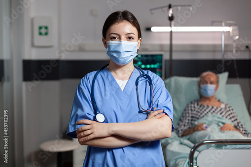 Portrait of medical nurse with chirurgical mask in hospital room with sick unweel senior pantient with trauma looking at camea with arms crossed, wearing scrubs.