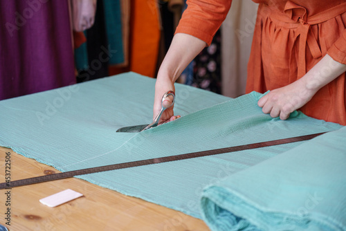 Cropped shot of woman tailor cutting fabric on table, working on new clothing at workplace in atelier or fashion studio with fashionable dresses hanging on clothes rack stand on background.