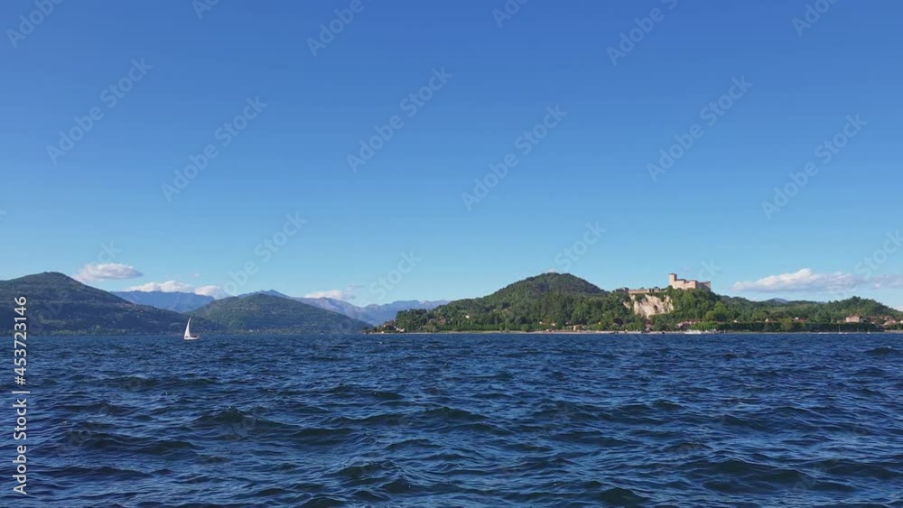 Small sailboat sailing on Maggiore lake agitated waters with mountains and Angera castle in background, Italy Small sailboat sailing on Maggiore lake agitated waters with mountains and Angera castle in background, Italy