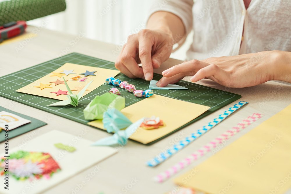 Close-up image of woman making elements like stars, crane or flowers for greeting cards out of colored paper Close-up image of woman making elements like stars, crane or flowers for greeting cards out of colored paper