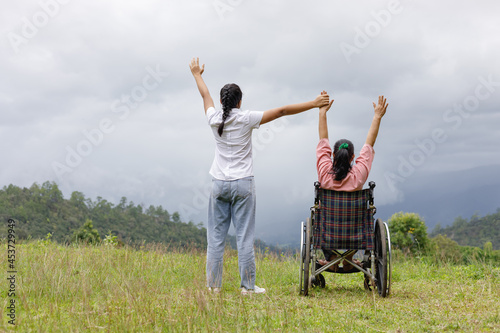 Disabled handicapped woman in wheelchair and care helper raised hands on mountain meadow park in sunny day. International Disability Day concept.