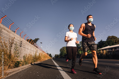 Sportive Asian man and woman in face mask running on street together during Coronavirus or Covid-19 outbreak. Runner couple jogging during quarantine. wide angle