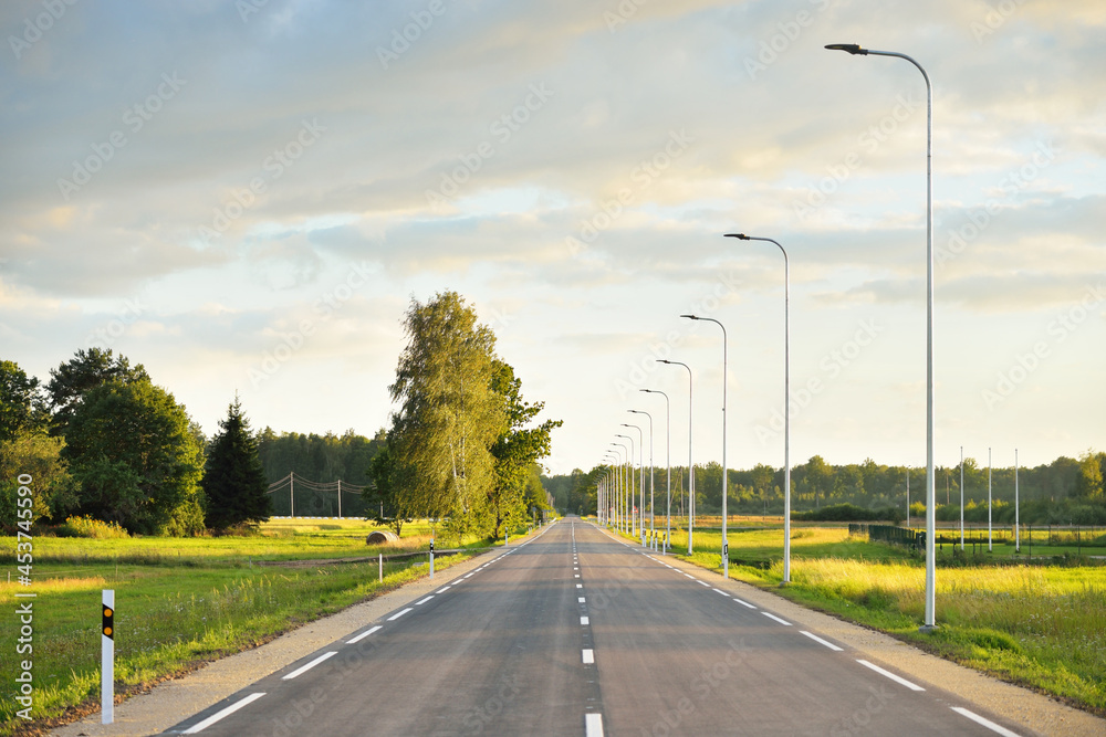 An empty highway (new asphalt road) through the green country fields and birch forest at sunset. Street lights close-up. Dramatic glowing clouds. Travel destinations, vacations, driving, freedom An empty highway (new asphalt road) through the green country fields and birch forest at sunset. Street lights close-up. Dramatic glowing clouds. Travel destinations, vacations, driving, freedom