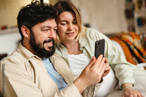 White couple smiling and using cellphone while sitting on couch at home