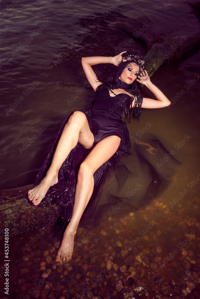 Sensual Beautiful Brunette Female In Black Dress Posing At Sea On Tree While Wearing Seashell Decorated Crown And Long Black Pearl Necklace. Sensual Beautiful Brunette Female In Black Dress Posing At Sea On Tree While Wearing Seashell Decorated Crown And Long Black Pearl Necklace.