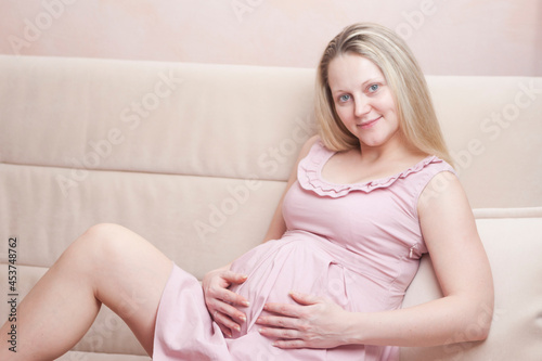 Young Romantic Caucasian Pregnant Blond Female Resting on Couch With Hands on Belly Posing Indoors.