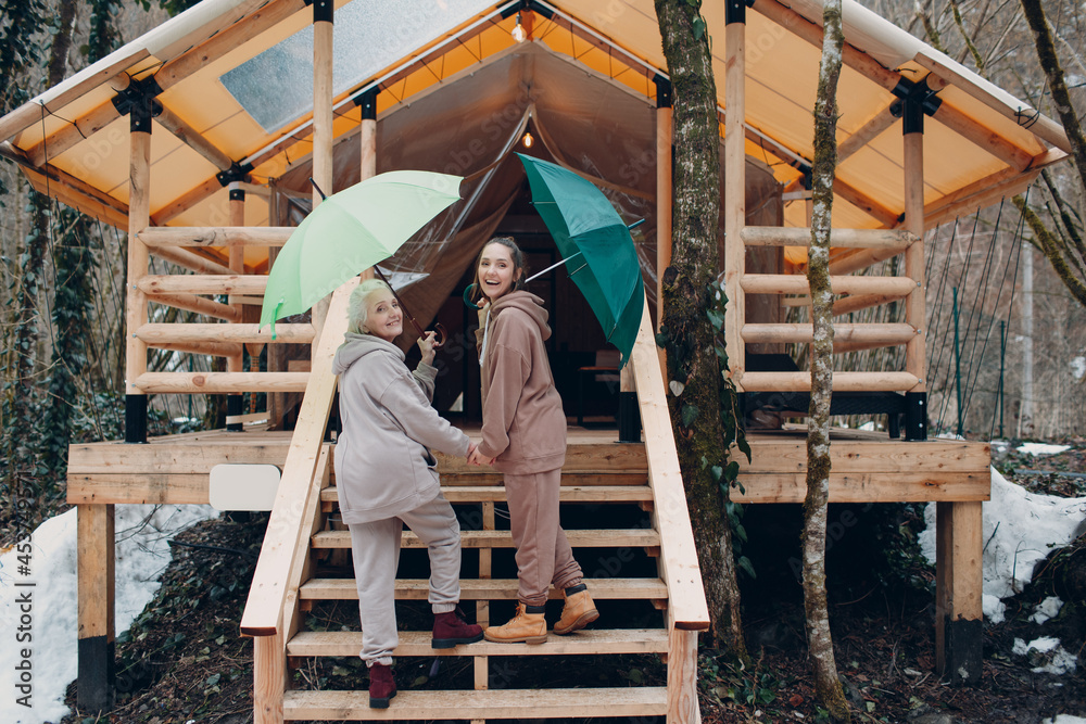 Elderly and young adult women with umbrella at glamping camping tent. Modern vacation lifestyle concept