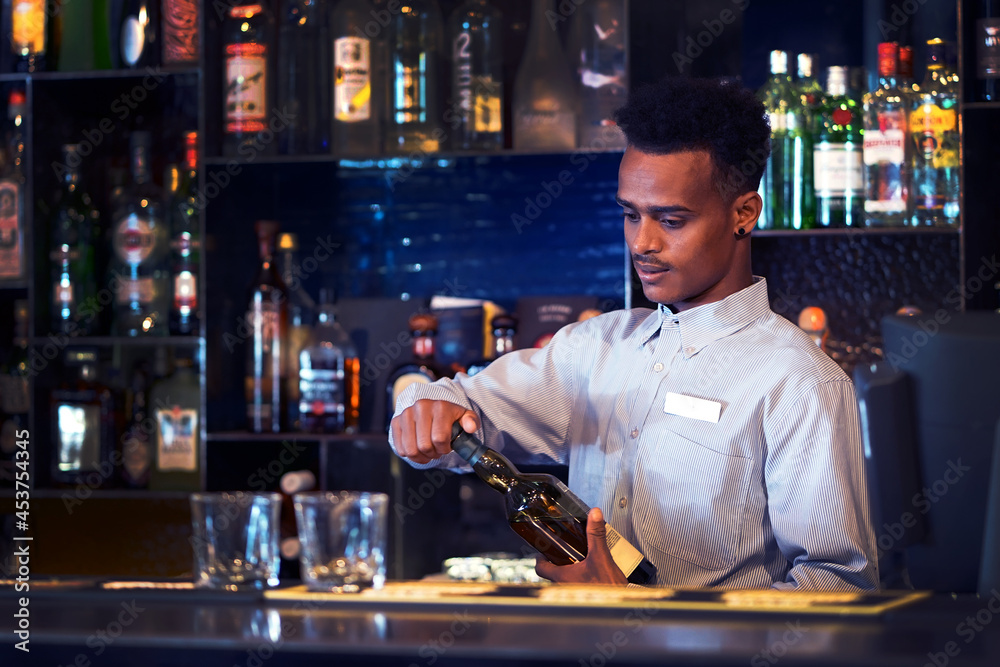 African young bartender opens a bottle of whiskey. Shelves with bottles of alcohol in the background.