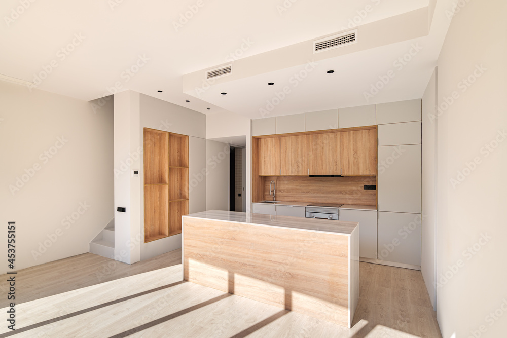 Fototapeta premium Empty interior of refurbished apartment with natural sunlight shining through big windows. Modern kitchen with island and wooden furniture.