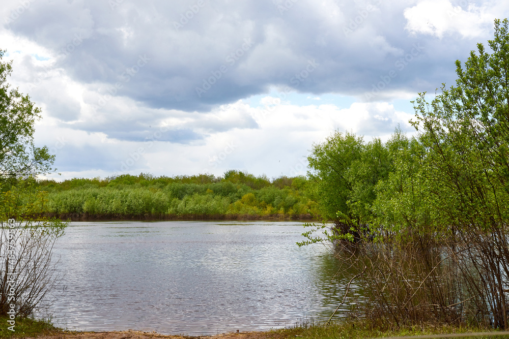 View of the water of a river or lake and the shore with grass and trees on a cloudy spring day