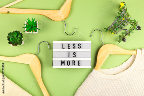 Less is more - written on lightbox next to Shopping cart entwined with plants on green background among wooden hangers. Conscious consumption slow fashion Zero waste concept. Top view flat lay photo