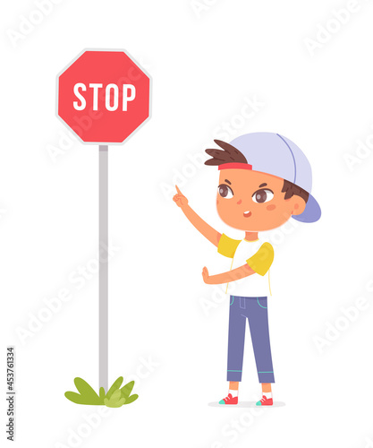 Boy in shirt with short sleeves and a blue baseball cap stands near a roadway on the sidewalk, and shows his hand on a road sign stop sign warns drivers of vehicles to be careful