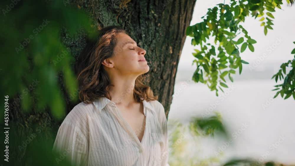 A young adult woman standing under the crown of a tree leaning on a trunk and enjoys the view and fresh air. A young adult woman standing under the crown of a tree leaning on a trunk and enjoys the view and fresh air.