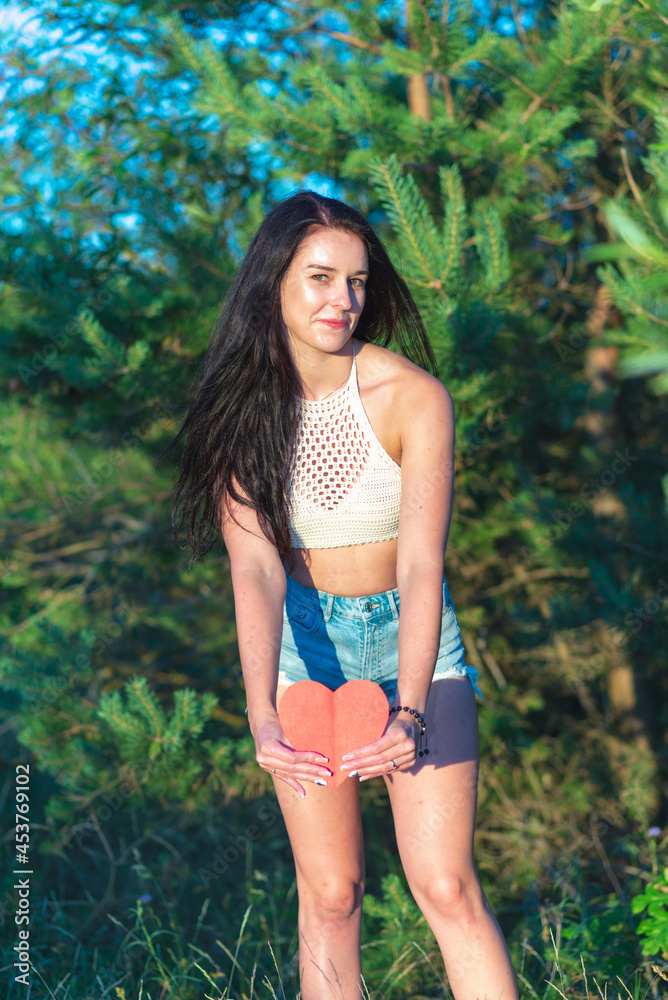 Pretty brunette showing red heart smiling dreamy nature summer sunny day.Woman in nature holds red paper heart in front Pretty brunette showing red heart smiling dreamy nature summer sunny day.Woman in nature holds red paper heart in front