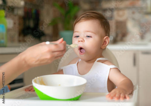 Feeding child. Cute baby eating food. His mother feeds him with a spoon. First lure. Portrait of a happy young child in a high chair being fed.