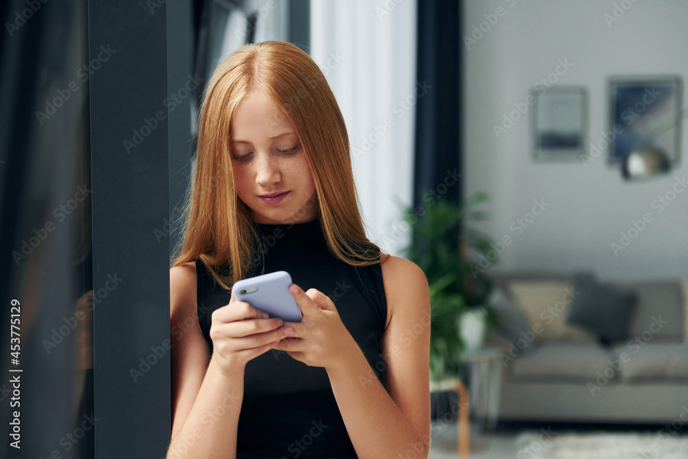 Standing with phone. Female teenager with blonde hair is at home at daytime Standing with phone. Female teenager with blonde hair is at home at daytime