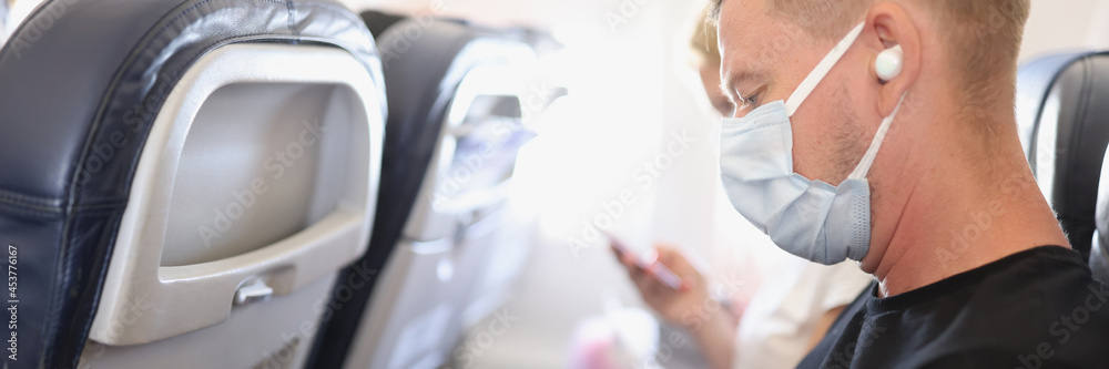 Obraz premium Man in medical protective mask sits on plane and watches video on his smartphone
