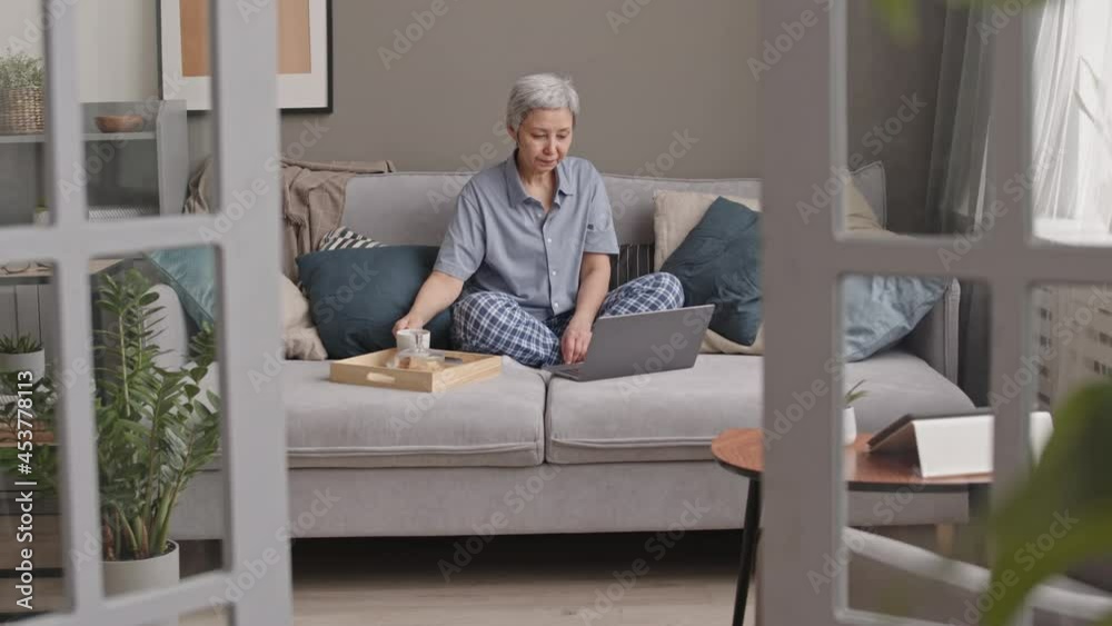 Tracking right of senior Asian woman wearing loungewear, sitting with crossed legs up on couch at home, using laptop computer