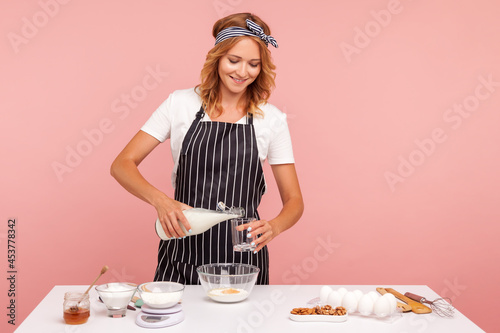 Photography Young adult woman chef pours the milk into glass, confectioner in black apron kneads the dough, preparing cakes or cookies, homemade pastry