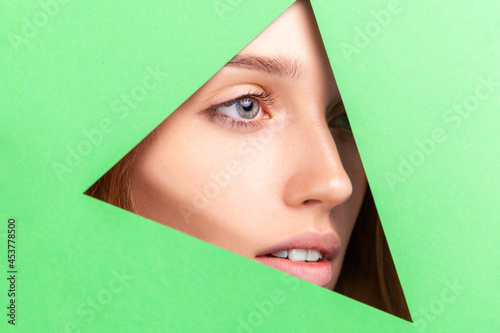 Face of young beautiful dreamy girl with perfect skin and without makeup, looking away across triangular hole in green paper. Beauty concept. Indoor studio shot. © khosrork