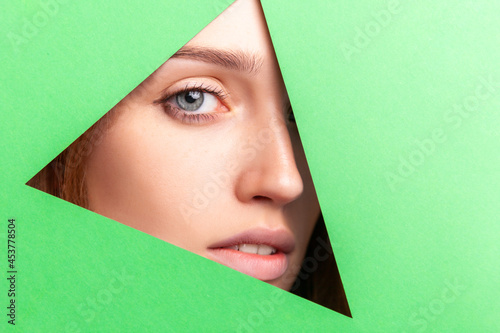 Face of attractive young female without makeup, woman looking at camera, looking through triangular, hole of green paper. Skin care and beauty concept. Indoor studio shot. © khosrork
