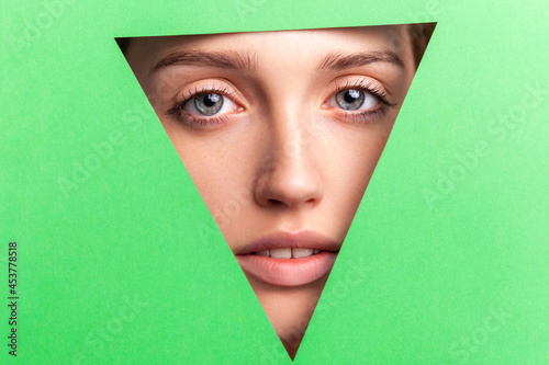 Closeup portrait of well-groomed woman with natural makeup, looking at camera through triangular hole of green paper, beauty and health and skin care concept. Indoor studio shot. © khosrork