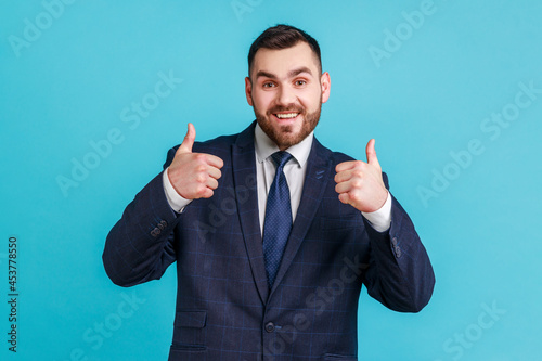 Handsome man with beard wearing official style suit looking at camera with toothy smile, showing thumbs up, being in good mood, hears excellent news. Indoor studio shot isolated on blue background.