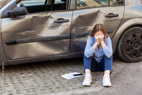 Portrait of blonde female wearing casual attire sitting on the ground and covering face with palms, being in despair after road accident, damaged side part of vehicle and doors. Outdoor shot. © khosrork
