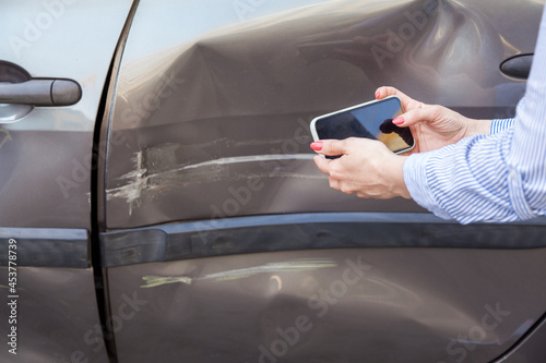 Unknown woman with mobile phone in hands taking picture of damaged part of car, photographing scratches and dents on auto door, records the consequences of the accident. Outdoor shot.