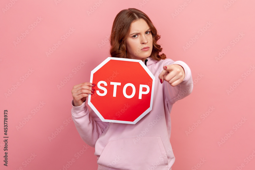 Portrait of serious curly haired teenage girl in hoodie holding big red stop road sign and pointing finger on you, strictly looking at camera. Indoor studio shot isolated on pink background Portrait of serious curly haired teenage girl in hoodie holding big red stop road sign and pointing finger on you, strictly looking at camera. Indoor studio shot isolated on pink background