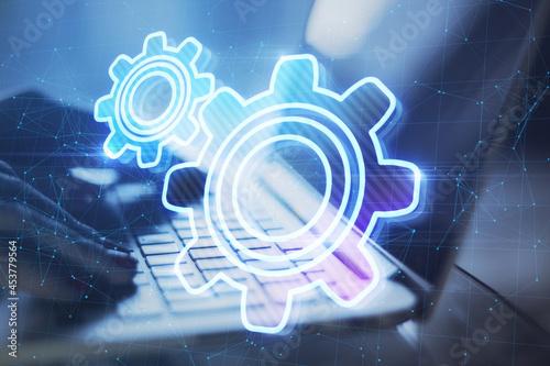 Close up of backlit hands typing on laptop keyboard at desktop with abstract glowing gear wheels on blurry background with connections. Digital engineering and industry concept. Double exposure.