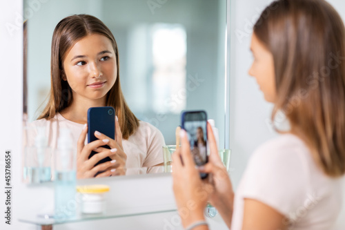 beauty, hygiene and people concept - teenage girl looking in mirror and taking selfie with smartphone at bathroom