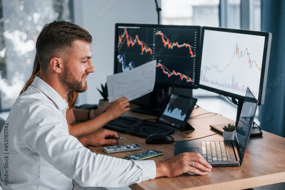 With woman. Young businessman in formal clothes is in office with multiple screens With woman. Young businessman in formal clothes is in office with multiple screens