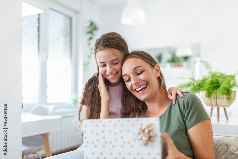 Young woman and girl at home celebrating mother's day sitting on sofa daughter hugging mother kissing cheek mom laughing joyful holding gift box Young woman and girl at home celebrating mother's day sitting on sofa daughter hugging mother kissing cheek mom laughing joyful holding gift box