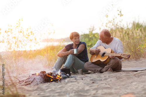 Senior couple Camping holiday in the summer nature. Camping concept. senior man playing guitar and singing song to his wife sitting on the beach near the fire
