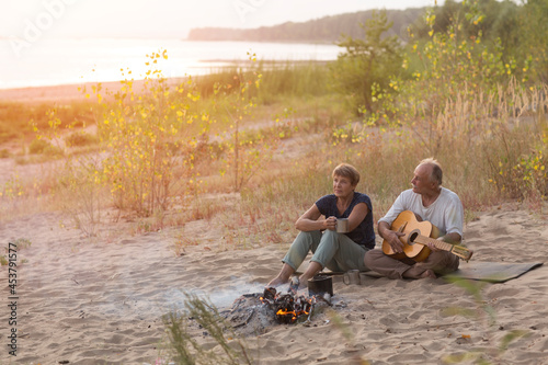 Senior couple Camping holiday in the summer nature. Camping concept. senior man playing guitar and singing song to his wife sitting on the beach near the fire 